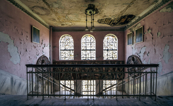 Ruined historic hall with arched windows, peeling pink walls, ornate ceiling, abandoned mansion, heritage decay, gothic restoration concept.