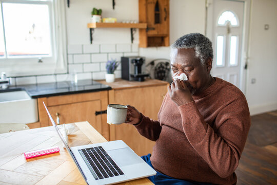 Senior man blowing nose while working from home in kitchen