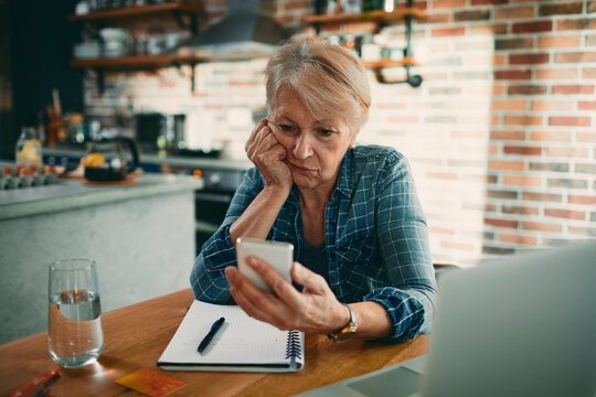 Senior woman worried about finances while using smartphone at kitchen table
