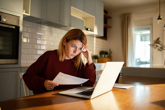 Worried woman checking household bills on laptop at kitchen table
