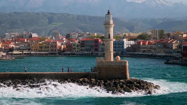 Greece Crete Chania 4K Drone Video Tracking Venetian Harbor with Lighthouse Crashing Waves and Distant Mountains over Old Town Scenic Marina and Fortress Travel