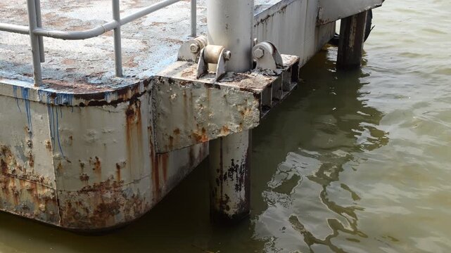 Close-up shot of rusty metal floating pier oscillating on murky water of Chao Phraya River. Camera focuses on pile bracket equipped with four-roller guide system that secures pier to vertical beam