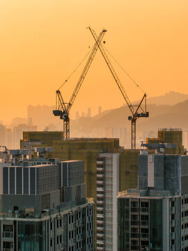 Towering construction cranes form a silhouette against a golden sunset sky over the dense urban skyline of Hong Kong representing growth and progress.