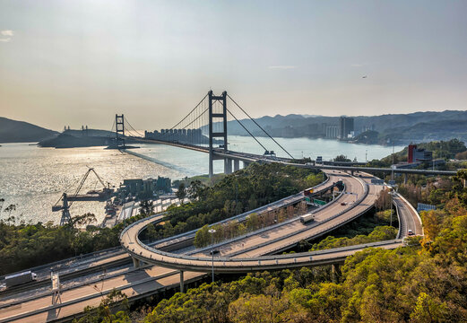 The Tsing Ma Bridge spans the water at sunset, connecting to a complex highway interchange in Hong Kong.