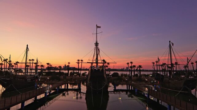Silhouette of the Santa Maria, Pinta, and Nina at a tropical pier during a vibrant purple and orange sunset.
