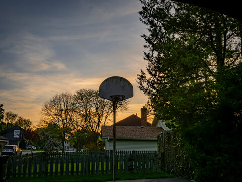 Suburban neighborhood scene at sunset in Naperville Illinois with a weathered basketball hoop backboard and fence in silhouette against a colorful sky