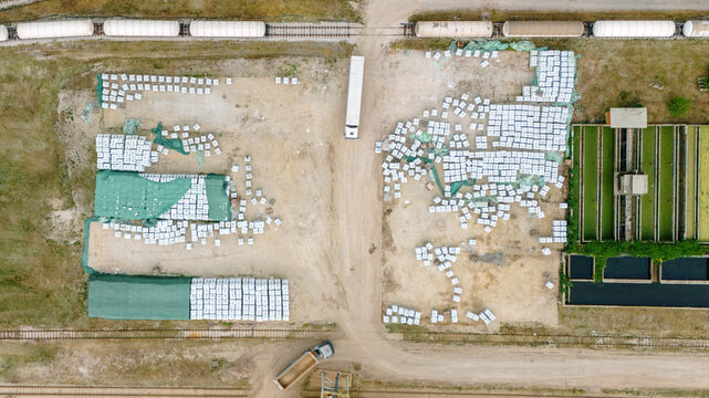 Aerial view of industrial storage yard with stacks of white bags, green tarps, a parked truck, and railway tracks with tanker cars in Sabac, Vojvodina, Serbia.