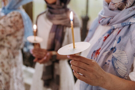 Women with covered heads holding lit wax candles during a religious ceremony inside an Orthodox church. Soft warm light and blurred background.