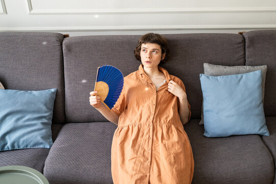 Woman on couch with sick expression waving hand paper fan overwhelmed by summer heat in apartment without air conditioning, annoyed female with fan in hand touches collar and looks with frustration.