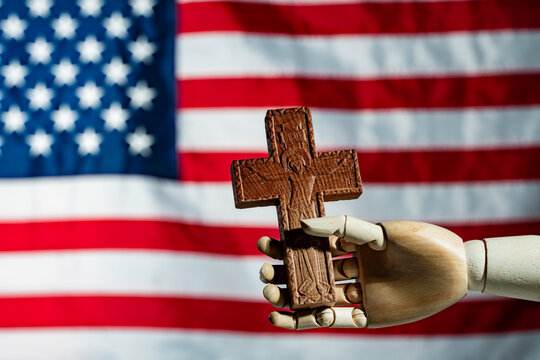 Wooden hand holding Christian cross in front of US flag. Mannequin hand holding carved wooden cross on American flag background. Concept of religion faith and Christian values in the United States