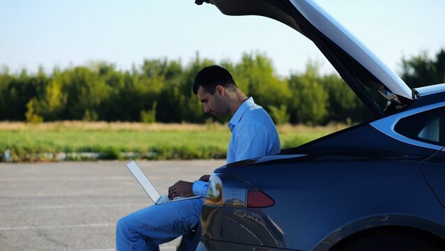 Young handsome entrepreneur works on notebook sitting in electric car trunk. Successful businessman in formal wear using computer at auto. Confident manager typing text on laptop outdoor. Slow mo