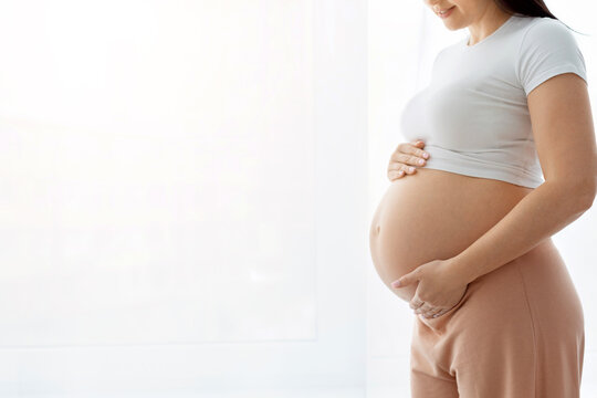 Closeup side profile of pregnant woman holding bare round belly with both hands near bright window, head cropped, wide copy space on left for text or graphic overlay