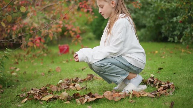 Girl crouching in leaf circle on grass, white sweater and jeans, braided hair, gently picking up brown leaves, garden backdrop with red shrub, soft autumn light, curious playful mood, seasonal