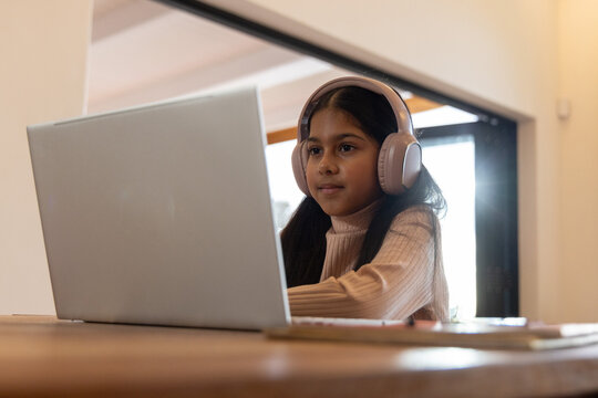 Indian female child sitting at wood table wearing ribbed sweater and headset using laptop by window