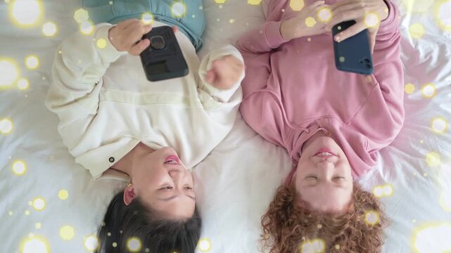Left woman angling phone, women on bed sharing screen and smiling, sparkle overlay accenting tech