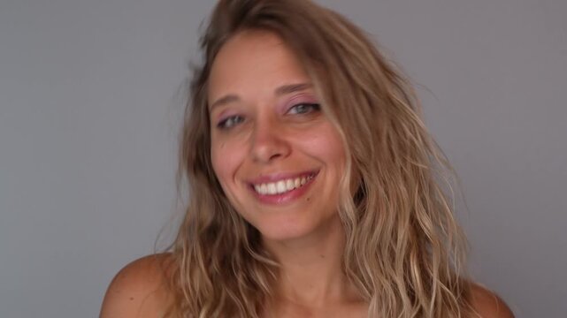 Young blonde woman playfully tousling wavy hair, smiling at camera. Positive mood, light flirt, natural volume and texture, carefree beauty and hair styling concept