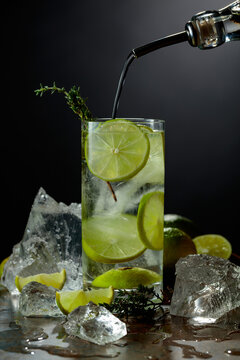 Gin is poured from a bottle into a glass with ice and lime slices.