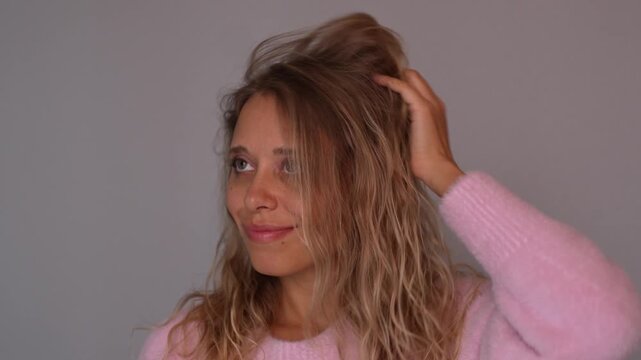 Young blonde woman lifting wavy hair styled with curly method, showing natural volume and texture. Soft pink cozy sweater, healthy haircare routine and modern beauty concept