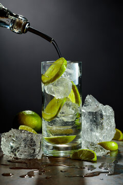 Gin is poured from a bottle into a glass with ice and lime slices.
