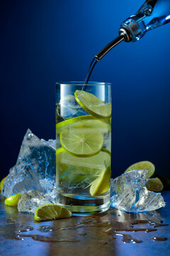 Gin is poured from a bottle into a glass with lime slices and crushed ice.