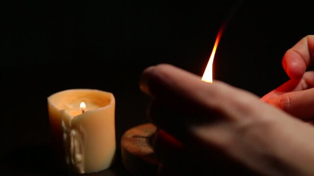 A woman lights a cone-shaped aroma incense with a match, placed in a vintage metal box on a wooden stand, while a candle burns on a table. She then closes the box with a lid, and smoke rises from it