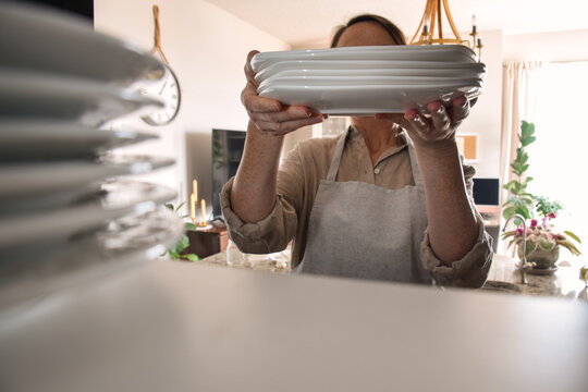 Woman placing stack of clean white plates into kitchen cupboard, viewed from inside the cabinet. Point of view perspective highlighting organization, storage, and tidy home routine in modern kitchen