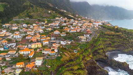 A high-angle aerial view of a coastal town on Madeira island, featuring orange-roofed houses, terraced green hillsides, and steep cliffs overlooking the Atlantic Ocean. © JULIA