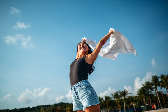 Happy woman celebrating freedom holding white shirt in the wind against blue sky at tropical beach