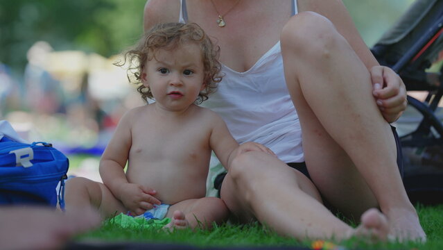 Toddler holding colorful toy before throwing it on grass and standing up near mother during summer picnic, playful outdoor childhood moment filled with curiosity and energy