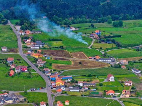 Aerial view of a rural village with colorful houses, green agricultural fields, and smoke rising from a fire near Cudillero, Asturias, Spain.