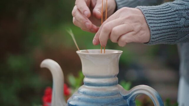 Closeup hands arranging dried stems in blue teapot, home stylist places grasses into ceramic vessel on lacecovered table, cozy sweater sleeves, slow careful motion, rustic centerpiece preparation