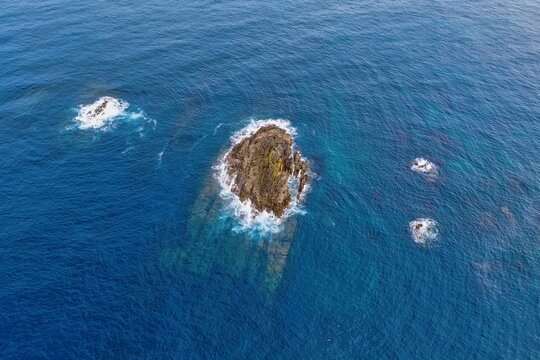 Aerial view of rocky islets and stacks surrounded by deep blue ocean water and white foam near Silence Beach Viewpoint in Cudillero, Asturias, Spain.