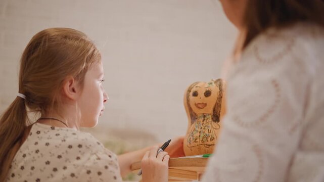 Young girl painting pumpkin with mother in sunlit kitchen, warm tones, drawing friendly face on small pumpkin on wooden table, markers and brushes nearby, adult guiding, playful autumn craft, family