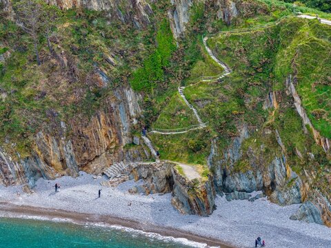 Aerial view of Silence Beach Viewpoint with a winding stone staircase on steep cliffs and people walking down to the pebble beach, Cudillero, Asturias, Spain.