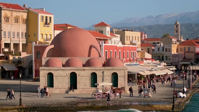 Chania Crete Aerial 4K. Pink Mosque Giali Tzamisi and horse carriages on promenade. Drone flyover to old town church bell tower. White Mountains landscape. April spring travel Greece heritage scenery