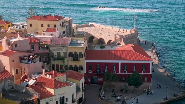 Chania Old Harbor Crete. Aerial 4K. Drone reveal from pastel Venetian buildings to lighthouse with crashing waves. Chania promenade with people walking. Greece spring travel cinematic