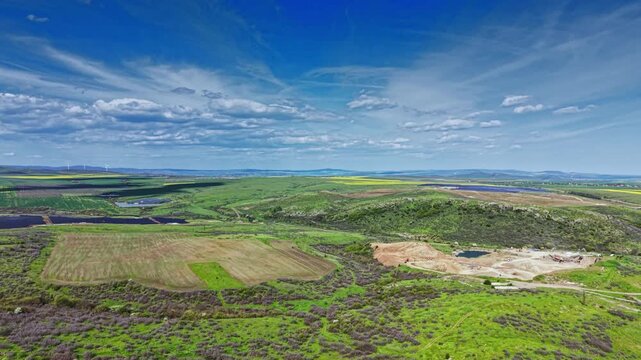 In the late afternoon, green fields stretch across the rural landscape with patches of cultivated land. Distant hills and a blue sky with clouds create a vast view.