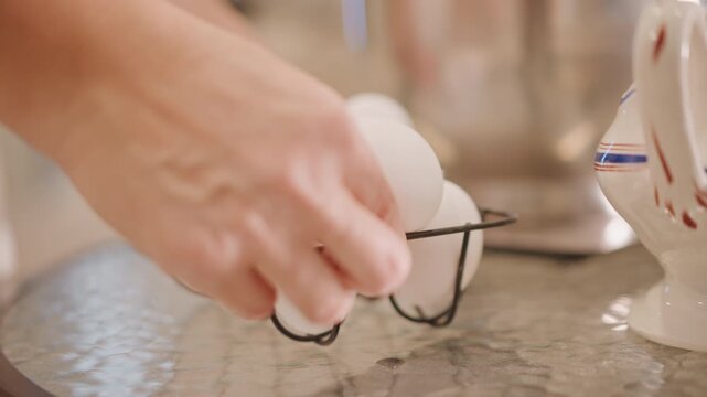 Closeup hand placing eggs in basket, home baker gently sets white eggs into vintage wire basket on glass table, ceramic pitcher and mixer blurred in background, warm soft light, preparing ingredients