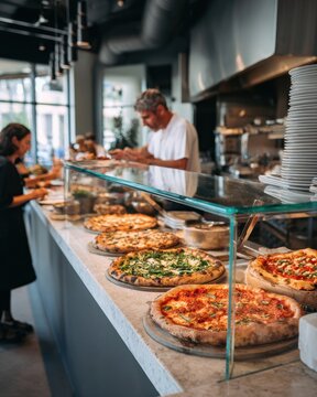Pizzeria counter with display of fresh pizzas, glass case, customers ordering