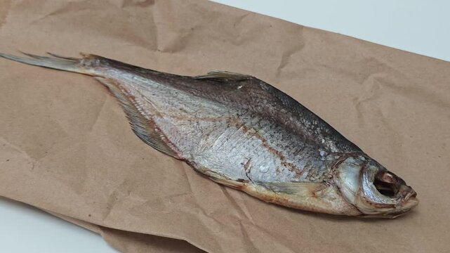 A single, uncleaned, dry-cured fish, typically enjoyed as a salty snack with beer, is presented on rustic brown packaging paper, rotating against a simple white backdrop, highlighting its texture and