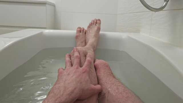 A relaxing bath scene from a first-person view, featuring a man's legs and hands gently moving through clear water in a white bathtub, providing a soothing and peaceful personal care moment at home.