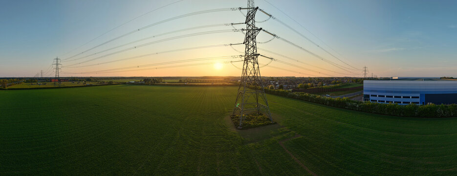 Aerial view of high voltage electricity pylons and power lines stretching across green agricultural fields and industrial warehouses during a golden sunset Armthorpe, England, United Kingdom.