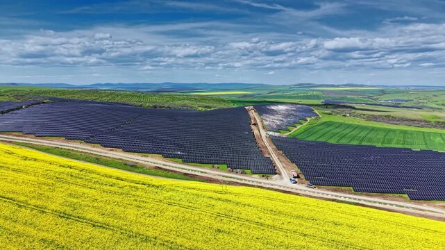 Workers are installing solar panels in a vast field surrounded by green land and an open sky. The site shows rows of solar panels arranged in a systematic manner.