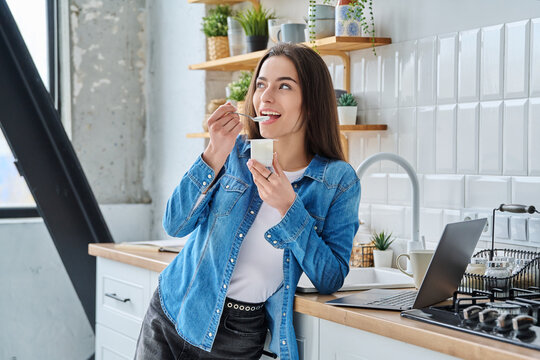 Attractive young woman in denim shirt holding spoon and eating white yogurt, in modern kitchen