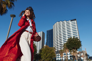 Obraz premium Cheerful woman outdoors wearing a red coat, walking under a bright blue sky, with modern city towers in the background, catching a warm sunlit moment and a carefree smile today