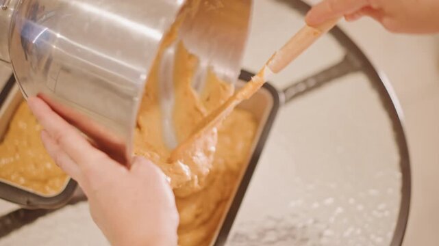Scraping pumpkin batter into loaf pan closeup of spatula scraping bowl to release glossy batter, tilting stainless mixing bowl, smoothing top for even bake, warm kitchen ambiance and aromatic spices