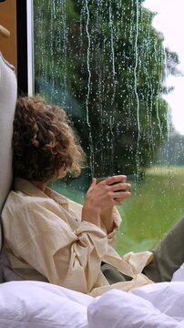 Pensive young woman with curly hair sitting on a cozy bed with a warm drink while watching the pouring rain outside the window, enjoying a moment of tranquility and relaxation at home