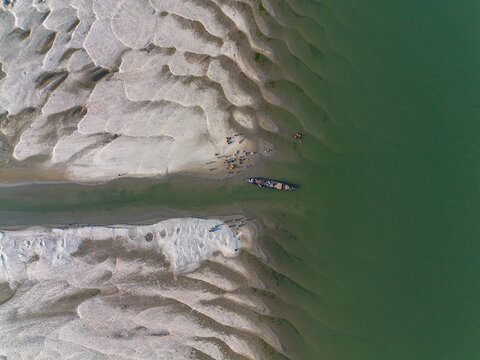 Aerial view of a traditional wooden boat with people navigating through a narrow channel between textured sandbars in Gaibandha, Rangpur Division, Bangladesh.