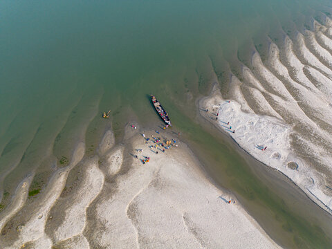 Aerial view of a wooden boat docked on a sandy riverbank with people gathered near the water and ripple patterns in the sand in Gaibandha, Rangpur Division, Bangladesh.