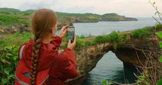 Traveler woman takes photo using smartphone majestic Broken Beach natural rock arch Nusa Penida Bali Indonesia